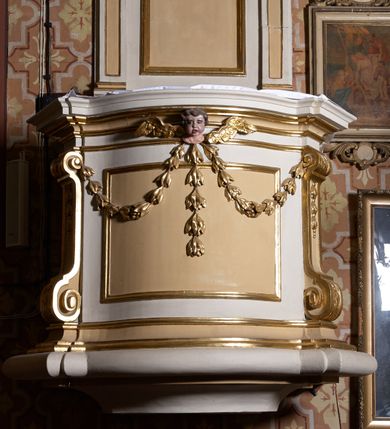 Zdjęcie nr 2: Ambona przyścienna, wisząca. Kosz czworoboczny, wybrzuszony z przodu na rzucie łuku odcinkowego. Na narożach flankowany pilastrami, zdobionymi wolutami. Ścianki korpusu dekorowane płycinami w kształcie leżącego prostokąta. Parapet w formie profilowanego gzymsu. Na ściance przedniej, w partii gzymsu, uskrzydlona główka aniołka, spod której zwisa girlanda utworzona z kampanul. Kosz ambony ścięty od dołu prosto. Zaplecek ujęty parą pilastrów, z płycinami zdobionymi w części górnej zwisami ze stylizowanych kampanul, z nałożonymi u szczytu konsolkami, pełniącymi funkcję kapiteli, które wspierają odcinki przerwanego belkowania. Pośrodku zaplecka płycina w kształcie stojącego prostokąta zamknięta od góry łukiem wklęsłym, nad nią stylizowana muszla palmetowa, poniżej zwis utworzony z gałązki róży. Zwieńczenie w formie promienistej glorii, usytuowanej pomiędzy dwiema wolutami osadzonymi na odcinkach belkowania na osiach pilastrów. Gloria utworzona z rozszerzających się wiązek promieni o nierównej długości, pośrodku gołębica Ducha Świętego w otoku chmur. Struktura ambony malowana w kolorze białym, płyciny i detale w kolorze beżowo-kremowym, ornamentyka i profilowania złocone i srebrzone.