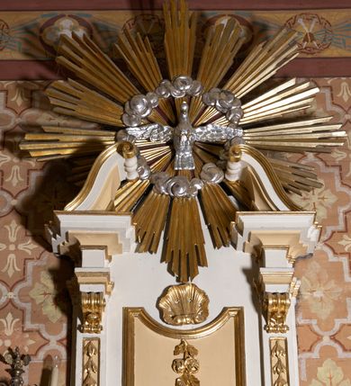 Zdjęcie nr 3: Ambona przyścienna, wisząca. Kosz czworoboczny, wybrzuszony z przodu na rzucie łuku odcinkowego. Na narożach flankowany pilastrami, zdobionymi wolutami. Ścianki korpusu dekorowane płycinami w kształcie leżącego prostokąta. Parapet w formie profilowanego gzymsu. Na ściance przedniej, w partii gzymsu, uskrzydlona główka aniołka, spod której zwisa girlanda utworzona z kampanul. Kosz ambony ścięty od dołu prosto. Zaplecek ujęty parą pilastrów, z płycinami zdobionymi w części górnej zwisami ze stylizowanych kampanul, z nałożonymi u szczytu konsolkami, pełniącymi funkcję kapiteli, które wspierają odcinki przerwanego belkowania. Pośrodku zaplecka płycina w kształcie stojącego prostokąta zamknięta od góry łukiem wklęsłym, nad nią stylizowana muszla palmetowa, poniżej zwis utworzony z gałązki róży. Zwieńczenie w formie promienistej glorii, usytuowanej pomiędzy dwiema wolutami osadzonymi na odcinkach belkowania na osiach pilastrów. Gloria utworzona z rozszerzających się wiązek promieni o nierównej długości, pośrodku gołębica Ducha Świętego w otoku chmur. Struktura ambony malowana w kolorze białym, płyciny i detale w kolorze beżowo-kremowym, ornamentyka i profilowania złocone i srebrzone.