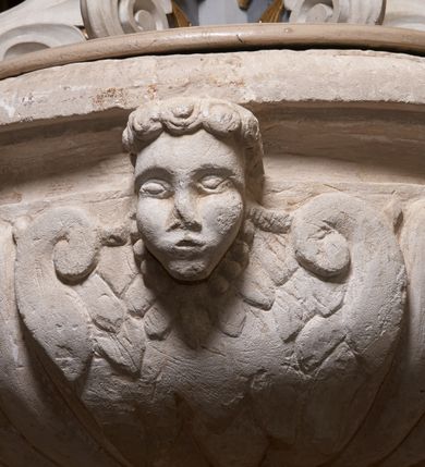 Zdjęcie nr 3: Chrzcielnica ustawiona na zdwojonym cokole, na planie kwadratu, o dolnej części w formie wysokiej, prostopadłościennej płyty i górnej, zwężającej się, o bocznych powierzchniach ugiętych, zarysowanych na linii łuków odcinkowych, dekorowanej liśćmi akantu. Trzon w formie kwadratowego w rzucie, gładkiego filaru podtrzymujący półkolistą, spłaszczoną czarę. Czara zdobiona od dołu puklowaniem oraz czterema, uskrzydlonymi główkami aniołków. Pokrywa w formie dwustopniowego postumentu złożonego z dwóch płyt połączonych akantowymi wolutami. Na górnym postumencie umieszczona  grupa rzeźbiarska przedstawiająca Chrzest Chrystusa. Chrystus ukazany w pozycji klęczącej, odziany w białe perizonium, prawą ręką czyniący gest błogosławieństwa, lewą trzymający ugiętą w łokciu. Twarz pociągła, z długim i wąskim nosem, wzrokiem skierowanym na wprost, okolona krótką brodą, zakończoną w dwa pukle. Włosy długie, kręcone opadające na plecy i ramiona. Święty Jan Chrzciciel ukazany w postawie stojącej, zwrócony w trzech czwartych w stronę Chrystusa, z lewą nogą wysuniętą do przodu, prawą ręką polewający głowę Jezusa wodą z muszli, zaś lewą przytrzymujący go za nadgarstek lewej dłoni. Święty ubrany w krótką szatę ze skóry wielbłądziej oraz czerwony płaszcz zarzucony na prawe ramię. Twarz pociągła, okolona średniej długości brodą, włosy krótkie, rozłożone w niedbałe pukle.  Włosy i zarost postaci – brązowe. Polichromia w odsłoniętych partiach karnacji ciała naturalistyczna. Sylwetki drobnej budowy, smukłe. Nad głową Chrystusa srebrzona gołębica przedstawiająca Ducha Świętego zatknięta na drucie.