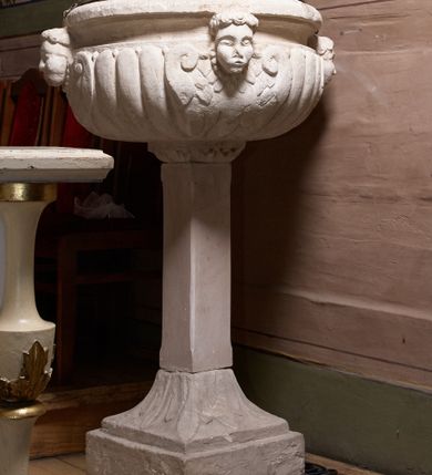 Zdjęcie nr 1: Chrzcielnica ustawiona na zdwojonym cokole, na planie kwadratu, o dolnej części w formie wysokiej, prostopadłościennej płyty i górnej, zwężającej się, o bocznych powierzchniach ugiętych, zarysowanych na linii łuków odcinkowych, dekorowanej liśćmi akantu. Trzon w formie kwadratowego w rzucie, gładkiego filaru podtrzymujący półkolistą, spłaszczoną czarę. Czara zdobiona od dołu puklowaniem oraz czterema, uskrzydlonymi główkami aniołków. Pokrywa w formie dwustopniowego postumentu złożonego z dwóch płyt połączonych akantowymi wolutami. Na górnym postumencie umieszczona  grupa rzeźbiarska przedstawiająca Chrzest Chrystusa. Chrystus ukazany w pozycji klęczącej, odziany w białe perizonium, prawą ręką czyniący gest błogosławieństwa, lewą trzymający ugiętą w łokciu. Twarz pociągła, z długim i wąskim nosem, wzrokiem skierowanym na wprost, okolona krótką brodą, zakończoną w dwa pukle. Włosy długie, kręcone opadające na plecy i ramiona. Święty Jan Chrzciciel ukazany w postawie stojącej, zwrócony w trzech czwartych w stronę Chrystusa, z lewą nogą wysuniętą do przodu, prawą ręką polewający głowę Jezusa wodą z muszli, zaś lewą przytrzymujący go za nadgarstek lewej dłoni. Święty ubrany w krótką szatę ze skóry wielbłądziej oraz czerwony płaszcz zarzucony na prawe ramię. Twarz pociągła, okolona średniej długości brodą, włosy krótkie, rozłożone w niedbałe pukle.  Włosy i zarost postaci – brązowe. Polichromia w odsłoniętych partiach karnacji ciała naturalistyczna. Sylwetki drobnej budowy, smukłe. Nad głową Chrystusa srebrzona gołębica przedstawiająca Ducha Świętego zatknięta na drucie.