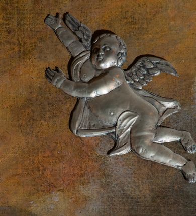 Zdjęcie nr 12: Obraz w kształcie stojącego prostokąta z przedstawieniem Wniebowzięcia Najświętszej Marii Panny. W centrum kompozycji ukazana Matka Boska unosząca się na chmurze obłoków, z ugiętymi w kolanach nogami, z prawą ręką wzniesioną do góry, lewą odsuniętą w bok, z głową lekko uniesioną do góry. Strój zasłonięty metalową, trybowaną sukienką. Twarz szeroka, z długim i wąskim nosem, wzrokiem skierowanym ku górze, włosy ciemne schowane pod metalową koroną; u szyi trzy sznury czerwonych korali. Wokół głowy nimb ze złotych gwiazd, a na głowie metalowa korona zamknięta. W dolnej części kompozycji grupka nagich putt w ekspresyjnych pozach, ukazane z różnych ujęć, z akcentem zielonej tkaniny, która przebiega pomiędzy nimi. Druga grupka putt nieco wyżej, po lewej stronie obrazu: jedno trzyma wianek, drugie zieloną gałązkę palmy. W górnej części obrazu, po bokach Marii, przybite dwa aniołki z blachy. Sukienka ozdobiona grawerowaną, bujną wicią roślinną z wielopłatkowymi kwiatami i liśćmi akantu. Tło ciemne w odcieniach szarości i brązu.

