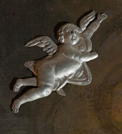 Zdjęcie nr 11: Obraz w kształcie stojącego prostokąta z przedstawieniem Wniebowzięcia Najświętszej Marii Panny. W centrum kompozycji ukazana Matka Boska unosząca się na chmurze obłoków, z ugiętymi w kolanach nogami, z prawą ręką wzniesioną do góry, lewą odsuniętą w bok, z głową lekko uniesioną do góry. Strój zasłonięty metalową, trybowaną sukienką. Twarz szeroka, z długim i wąskim nosem, wzrokiem skierowanym ku górze, włosy ciemne schowane pod metalową koroną; u szyi trzy sznury czerwonych korali. Wokół głowy nimb ze złotych gwiazd, a na głowie metalowa korona zamknięta. W dolnej części kompozycji grupka nagich putt w ekspresyjnych pozach, ukazane z różnych ujęć, z akcentem zielonej tkaniny, która przebiega pomiędzy nimi. Druga grupka putt nieco wyżej, po lewej stronie obrazu: jedno trzyma wianek, drugie zieloną gałązkę palmy. W górnej części obrazu, po bokach Marii, przybite dwa aniołki z blachy. Sukienka ozdobiona grawerowaną, bujną wicią roślinną z wielopłatkowymi kwiatami i liśćmi akantu. Tło ciemne w odcieniach szarości i brązu.

