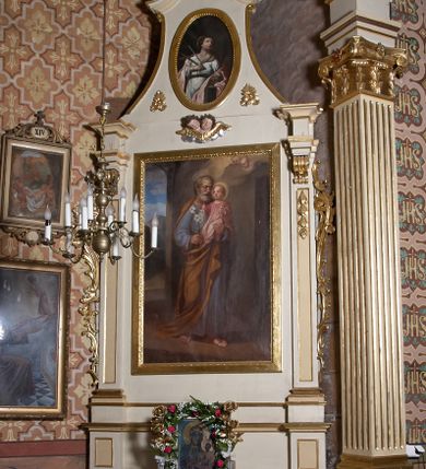 Zdjęcie nr 5: Obraz w kształcie stojącego prostokąta przedstawiający św. Józefa z Dzieciątkiem. Święty został ukazany w całej postaci, delikatnie zwrócony w lewo. Na lewej ręce trzyma Dzieciątko Jezus, a w prawej gałązkę białej lilii. Święty Józef ma szeroką twarz, okoloną krótką i siwą brodą oraz siwe włosy, nos długi i wąski, wzrok wpatrzony w Jezusa. Ubrany jest w niebieską szatę z długimi rękawami oraz ugrowy płaszcz; na stopach ma sandały. Dzieciątko ma twarz o rysach niemowlęcych, wzrok skierowany ku górze. Zwrócone jest w trzech czwartych w prawo, ukazane w pozycji siedzącej, z krzyżykiem łacińskim w lewej dłoni, z prawą rączką wyciągniętą przed siebie. Ubrane w jasnoróżową sukienkę z długimi rękawami. Wokół głów św. Józefa i Dzieciątka znajdują się złote nimby. Całość ukazana w sumarycznie oddanym wnętrzu architektonicznym z arkadą otwartą na pejzaż górski. W prawym górnym narożu dwie uskrzydlone główki anielskie na chmurze obłoków. 
