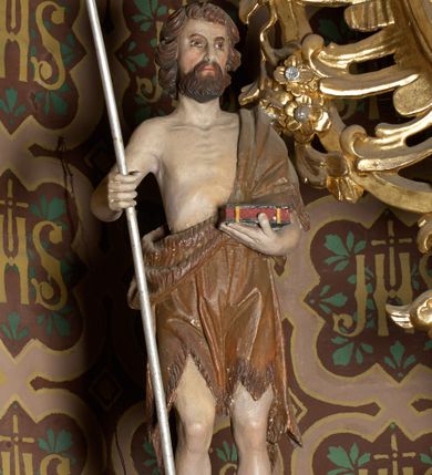Zdjęcie nr 3: Rzeźba wolnostojąca, przedstawia św. Jana Chrzciciela, ustawiona na niskim, zielonym cokole na planie kwadratu. Święty ukazany frontalnie, w postawie stojącej z krzyżem o długim drzewcu w prawej dłoni oraz zamkniętą księgą w lewej. Twarz podłużna z długim i wąskim nosem, ze wzrokiem skierowanym w dal, okolona brązową, średniej długości brodą oraz krótkimi włosami, zasłaniającymi uszy.  Święty jest ubrany w szatę uszytą ze skóry zwierzęcej, odsłaniającą nagi tors i nogi poniżej kolan. Sylwetka szczupła. Polichromia w odsłoniętych partiach ciała naturalistyczna. 