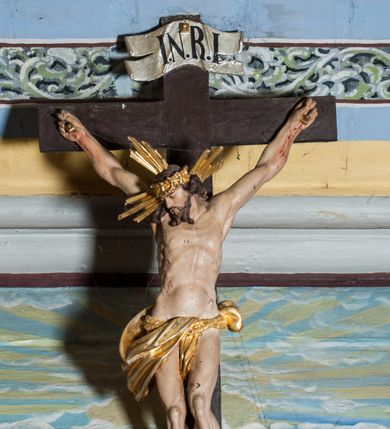 Zdjęcie nr 3: Krucyfiks zawieszony w łuku tęczowym kościoła. Do prostego krzyża przybita trzema gwoździami figura umarłego Chrystusa. Postać ukazana w dużym zwisie, o szeroko rozłożonych ramionach, z głową opadającą na prawy bark, nogi lekko ugięte w kolanach. Twarz Chrystusa jest podłużna, o zamkniętych oczach, długim i wąskim nosie, okolona jest krótką brodą rozdzieloną na końcu na dwa pukle i długimi włosami w postaci wijących się pukli, opadających na plecy i prawe ramię. Na głowie szeroka korona cierniowa, a wokół niej krzyżowy nimb promienisty. Ciało o wyraźnie podkreślonej anatomii, z silnie zaznaczoną muskulaturą. Perizonium przewieszone przez podwójny sznur, odsłaniające lewe biodro. Krzyż prosty, gładki, malowany na kolor ciemnobrązowy. Po lewej stronie krzyża nadlatujący aniołek, widoczny z lewego profilu, przepasany złotą tkaniną. Nad głową Chrystusa na pionowej belce banderola z napisem „I.N.R.I.” Polichromia w odsłoniętych partiach ciała naturalistyczna, szaty i detale złocone. 