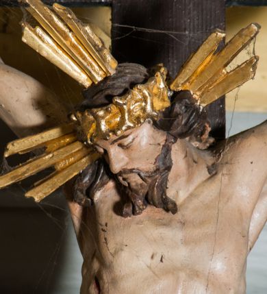 Zdjęcie nr 5: Krucyfiks zawieszony w łuku tęczowym kościoła. Do prostego krzyża przybita trzema gwoździami figura umarłego Chrystusa. Postać ukazana w dużym zwisie, o szeroko rozłożonych ramionach, z głową opadającą na prawy bark, nogi lekko ugięte w kolanach. Twarz Chrystusa jest podłużna, o zamkniętych oczach, długim i wąskim nosie, okolona jest krótką brodą rozdzieloną na końcu na dwa pukle i długimi włosami w postaci wijących się pukli, opadających na plecy i prawe ramię. Na głowie szeroka korona cierniowa, a wokół niej krzyżowy nimb promienisty. Ciało o wyraźnie podkreślonej anatomii, z silnie zaznaczoną muskulaturą. Perizonium przewieszone przez podwójny sznur, odsłaniające lewe biodro. Krzyż prosty, gładki, malowany na kolor ciemnobrązowy. Po lewej stronie krzyża nadlatujący aniołek, widoczny z lewego profilu, przepasany złotą tkaniną. Nad głową Chrystusa na pionowej belce banderola z napisem „I.N.R.I.” Polichromia w odsłoniętych partiach ciała naturalistyczna, szaty i detale złocone. 