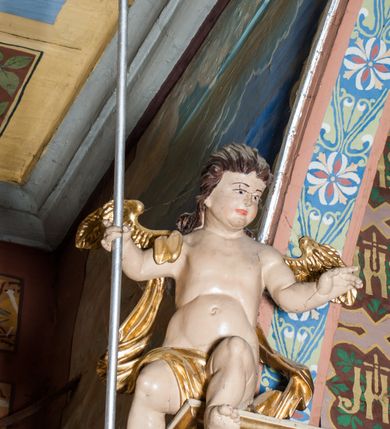 Zdjęcie nr 8: Krucyfiks zawieszony w łuku tęczowym kościoła. Do prostego krzyża przybita trzema gwoździami figura umarłego Chrystusa. Postać ukazana w dużym zwisie, o szeroko rozłożonych ramionach, z głową opadającą na prawy bark, nogi lekko ugięte w kolanach. Twarz Chrystusa jest podłużna, o zamkniętych oczach, długim i wąskim nosie, okolona jest krótką brodą rozdzieloną na końcu na dwa pukle i długimi włosami w postaci wijących się pukli, opadających na plecy i prawe ramię. Na głowie szeroka korona cierniowa, a wokół niej krzyżowy nimb promienisty. Ciało o wyraźnie podkreślonej anatomii, z silnie zaznaczoną muskulaturą. Perizonium przewieszone przez podwójny sznur, odsłaniające lewe biodro. Krzyż prosty, gładki, malowany na kolor ciemnobrązowy. Po lewej stronie krzyża nadlatujący aniołek, widoczny z lewego profilu, przepasany złotą tkaniną. Nad głową Chrystusa na pionowej belce banderola z napisem „I.N.R.I.” Polichromia w odsłoniętych partiach ciała naturalistyczna, szaty i detale złocone. 