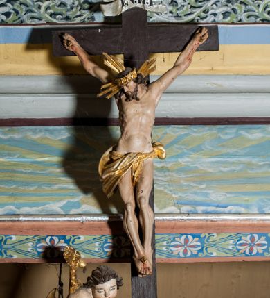 Zdjęcie nr 1: Krucyfiks zawieszony w łuku tęczowym kościoła. Do prostego krzyża przybita trzema gwoździami figura umarłego Chrystusa. Postać ukazana w dużym zwisie, o szeroko rozłożonych ramionach, z głową opadającą na prawy bark, nogi lekko ugięte w kolanach. Twarz Chrystusa jest podłużna, o zamkniętych oczach, długim i wąskim nosie, okolona jest krótką brodą rozdzieloną na końcu na dwa pukle i długimi włosami w postaci wijących się pukli, opadających na plecy i prawe ramię. Na głowie szeroka korona cierniowa, a wokół niej krzyżowy nimb promienisty. Ciało o wyraźnie podkreślonej anatomii, z silnie zaznaczoną muskulaturą. Perizonium przewieszone przez podwójny sznur, odsłaniające lewe biodro. Krzyż prosty, gładki, malowany na kolor ciemnobrązowy. Po lewej stronie krzyża nadlatujący aniołek, widoczny z lewego profilu, przepasany złotą tkaniną. Nad głową Chrystusa na pionowej belce banderola z napisem „I.N.R.I.” Polichromia w odsłoniętych partiach ciała naturalistyczna, szaty i detale złocone. 