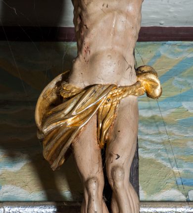 Zdjęcie nr 4: Krucyfiks zawieszony w łuku tęczowym kościoła. Do prostego krzyża przybita trzema gwoździami figura umarłego Chrystusa. Postać ukazana w dużym zwisie, o szeroko rozłożonych ramionach, z głową opadającą na prawy bark, nogi lekko ugięte w kolanach. Twarz Chrystusa jest podłużna, o zamkniętych oczach, długim i wąskim nosie, okolona jest krótką brodą rozdzieloną na końcu na dwa pukle i długimi włosami w postaci wijących się pukli, opadających na plecy i prawe ramię. Na głowie szeroka korona cierniowa, a wokół niej krzyżowy nimb promienisty. Ciało o wyraźnie podkreślonej anatomii, z silnie zaznaczoną muskulaturą. Perizonium przewieszone przez podwójny sznur, odsłaniające lewe biodro. Krzyż prosty, gładki, malowany na kolor ciemnobrązowy. Po lewej stronie krzyża nadlatujący aniołek, widoczny z lewego profilu, przepasany złotą tkaniną. Nad głową Chrystusa na pionowej belce banderola z napisem „I.N.R.I.” Polichromia w odsłoniętych partiach ciała naturalistyczna, szaty i detale złocone. 