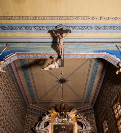 Zdjęcie nr 2: Krucyfiks zawieszony w łuku tęczowym kościoła. Do prostego krzyża przybita trzema gwoździami figura umarłego Chrystusa. Postać ukazana w dużym zwisie, o szeroko rozłożonych ramionach, z głową opadającą na prawy bark, nogi lekko ugięte w kolanach. Twarz Chrystusa jest podłużna, o zamkniętych oczach, długim i wąskim nosie, okolona jest krótką brodą rozdzieloną na końcu na dwa pukle i długimi włosami w postaci wijących się pukli, opadających na plecy i prawe ramię. Na głowie szeroka korona cierniowa, a wokół niej krzyżowy nimb promienisty. Ciało o wyraźnie podkreślonej anatomii, z silnie zaznaczoną muskulaturą. Perizonium przewieszone przez podwójny sznur, odsłaniające lewe biodro. Krzyż prosty, gładki, malowany na kolor ciemnobrązowy. Po lewej stronie krzyża nadlatujący aniołek, widoczny z lewego profilu, przepasany złotą tkaniną. Nad głową Chrystusa na pionowej belce banderola z napisem „I.N.R.I.” Polichromia w odsłoniętych partiach ciała naturalistyczna, szaty i detale złocone. 