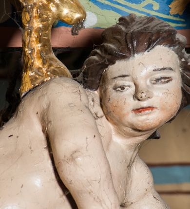 Zdjęcie nr 7: Krucyfiks zawieszony w łuku tęczowym kościoła. Do prostego krzyża przybita trzema gwoździami figura umarłego Chrystusa. Postać ukazana w dużym zwisie, o szeroko rozłożonych ramionach, z głową opadającą na prawy bark, nogi lekko ugięte w kolanach. Twarz Chrystusa jest podłużna, o zamkniętych oczach, długim i wąskim nosie, okolona jest krótką brodą rozdzieloną na końcu na dwa pukle i długimi włosami w postaci wijących się pukli, opadających na plecy i prawe ramię. Na głowie szeroka korona cierniowa, a wokół niej krzyżowy nimb promienisty. Ciało o wyraźnie podkreślonej anatomii, z silnie zaznaczoną muskulaturą. Perizonium przewieszone przez podwójny sznur, odsłaniające lewe biodro. Krzyż prosty, gładki, malowany na kolor ciemnobrązowy. Po lewej stronie krzyża nadlatujący aniołek, widoczny z lewego profilu, przepasany złotą tkaniną. Nad głową Chrystusa na pionowej belce banderola z napisem „I.N.R.I.” Polichromia w odsłoniętych partiach ciała naturalistyczna, szaty i detale złocone. 