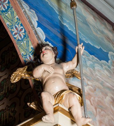 Zdjęcie nr 10: Krucyfiks zawieszony w łuku tęczowym kościoła. Do prostego krzyża przybita trzema gwoździami figura umarłego Chrystusa. Postać ukazana w dużym zwisie, o szeroko rozłożonych ramionach, z głową opadającą na prawy bark, nogi lekko ugięte w kolanach. Twarz Chrystusa jest podłużna, o zamkniętych oczach, długim i wąskim nosie, okolona jest krótką brodą rozdzieloną na końcu na dwa pukle i długimi włosami w postaci wijących się pukli, opadających na plecy i prawe ramię. Na głowie szeroka korona cierniowa, a wokół niej krzyżowy nimb promienisty. Ciało o wyraźnie podkreślonej anatomii, z silnie zaznaczoną muskulaturą. Perizonium przewieszone przez podwójny sznur, odsłaniające lewe biodro. Krzyż prosty, gładki, malowany na kolor ciemnobrązowy. Po lewej stronie krzyża nadlatujący aniołek, widoczny z lewego profilu, przepasany złotą tkaniną. Nad głową Chrystusa na pionowej belce banderola z napisem „I.N.R.I.” Polichromia w odsłoniętych partiach ciała naturalistyczna, szaty i detale złocone. 