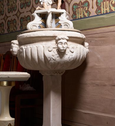 Zdjęcie nr 2: Pokrywa chrzcielnicy w formie figuralnej grupy rzeźbiarskiej przedstawiającej Chrzest Chrystusa. Rzeźby ustawione na postumencie skonstruowanym z użyciem sześciu liści stylizowanego akantu tworzących woluty łączące ze sobą dwie płyty: jedna dolna, większa, stanowi właściwą pokrywę zasłaniającą czarę chrzcielnicy, druga górna, znacznie mniejsza, pełni rolę postumentu pod rzeźby. Obie postaci ukazane w całości, pełnoplastyczne, przystosowane do oglądania ze wszystkich stron. Chrystus ukazany w pozycji klęczącej, prawą ręką czyniący gest błogosławieństwa, lewą trzymający ugiętą w łokciu. Twarz pociągła, z długim i wąskim nosem, wzrokiem skierowanym na wprost, okolona krótką brodą, zakończoną w dwa pukle. Włosy długie, kręcone opadające na plecy i ramiona; Chrystus odziany w białe perizonium. Święty Jan Chrzciciel ukazany w postawie stojącej, zwrócony w trzech czwartych w stronę Zbawiciela, z lewą nogą wysuniętą do przodu, prawą ręką polewający głowę Jezusa wodą z muszli, lewą zaś przytrzymujący go za nadgarstek lewej dłoni. Święty ubrany w krótką, brązową szatę ze skóry wielbłądziej oraz czerwony płaszcz zarzucony na prawe ramię. Twarz pociągła, okolona średniej długości brodą, włosy krótkie, rozłożone w niedbałe pukle. Polichromia w odsłoniętych partiach ciał naturalistyczna. Sylwetki drobnej budowy, smukłe. Nad głową Chrystusa gołębica Ducha Świętego, zamocowana na drucie. 