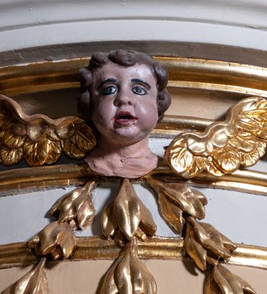 Zdjęcie nr 6: Pokrywa chrzcielnicy w formie figuralnej grupy rzeźbiarskiej przedstawiającej Chrzest Chrystusa. Rzeźby ustawione na postumencie skonstruowanym z użyciem sześciu liści stylizowanego akantu tworzących woluty łączące ze sobą dwie płyty: jedna dolna, większa, stanowi właściwą pokrywę zasłaniającą czarę chrzcielnicy, druga górna, znacznie mniejsza, pełni rolę postumentu pod rzeźby. Obie postaci ukazane w całości, pełnoplastyczne, przystosowane do oglądania ze wszystkich stron. Chrystus ukazany w pozycji klęczącej, prawą ręką czyniący gest błogosławieństwa, lewą trzymający ugiętą w łokciu. Twarz pociągła, z długim i wąskim nosem, wzrokiem skierowanym na wprost, okolona krótką brodą, zakończoną w dwa pukle. Włosy długie, kręcone opadające na plecy i ramiona; Chrystus odziany w białe perizonium. Święty Jan Chrzciciel ukazany w postawie stojącej, zwrócony w trzech czwartych w stronę Zbawiciela, z lewą nogą wysuniętą do przodu, prawą ręką polewający głowę Jezusa wodą z muszli, lewą zaś przytrzymujący go za nadgarstek lewej dłoni. Święty ubrany w krótką, brązową szatę ze skóry wielbłądziej oraz czerwony płaszcz zarzucony na prawe ramię. Twarz pociągła, okolona średniej długości brodą, włosy krótkie, rozłożone w niedbałe pukle. Polichromia w odsłoniętych partiach ciał naturalistyczna. Sylwetki drobnej budowy, smukłe. Nad głową Chrystusa gołębica Ducha Świętego, zamocowana na drucie. 