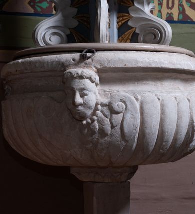 Zdjęcie nr 5: Pokrywa chrzcielnicy w formie figuralnej grupy rzeźbiarskiej przedstawiającej Chrzest Chrystusa. Rzeźby ustawione na postumencie skonstruowanym z użyciem sześciu liści stylizowanego akantu tworzących woluty łączące ze sobą dwie płyty: jedna dolna, większa, stanowi właściwą pokrywę zasłaniającą czarę chrzcielnicy, druga górna, znacznie mniejsza, pełni rolę postumentu pod rzeźby. Obie postaci ukazane w całości, pełnoplastyczne, przystosowane do oglądania ze wszystkich stron. Chrystus ukazany w pozycji klęczącej, prawą ręką czyniący gest błogosławieństwa, lewą trzymający ugiętą w łokciu. Twarz pociągła, z długim i wąskim nosem, wzrokiem skierowanym na wprost, okolona krótką brodą, zakończoną w dwa pukle. Włosy długie, kręcone opadające na plecy i ramiona; Chrystus odziany w białe perizonium. Święty Jan Chrzciciel ukazany w postawie stojącej, zwrócony w trzech czwartych w stronę Zbawiciela, z lewą nogą wysuniętą do przodu, prawą ręką polewający głowę Jezusa wodą z muszli, lewą zaś przytrzymujący go za nadgarstek lewej dłoni. Święty ubrany w krótką, brązową szatę ze skóry wielbłądziej oraz czerwony płaszcz zarzucony na prawe ramię. Twarz pociągła, okolona średniej długości brodą, włosy krótkie, rozłożone w niedbałe pukle. Polichromia w odsłoniętych partiach ciał naturalistyczna. Sylwetki drobnej budowy, smukłe. Nad głową Chrystusa gołębica Ducha Świętego, zamocowana na drucie. 