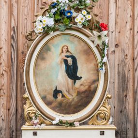 Zdjęcie nr 1: Feretron dwustronny z przedstawieniami Matki Boskiej Niepokalanie Poczętej oraz Matki Boskiej Szkaplerznej ustawiony na prostopadłościennym, profilowanym, zaopatrzonym w otwory na drążki do noszenia cokole. Obrazy w kształcie owalu, umieszczone w profilowanych ramach. Od dołu i góry zdobione liśćmi rzeźbionego akantu. Na obrazie Matki Boskiej Niepokalanie Poczętej Maria ukazana w centrum kompozycji na tle jasnoszarych chmur w postawie stojącej na półksiężycu, delikatnie zwrócona trzy czwarte w swoją prawą stronę. Ma dłonie złożone na piersi, głowę skierowaną w lewo. Ubrana jest w białą suknię z długimi rękawami, zasłaniającą stopy oraz niebieski płaszcz zarzucony jedną połą na lewe przedramię, drugi koniec płaszcza widoczny jest na dole, po prawej stronie Marii. Pod płaszczem chowa się małe putto. Twarz Marii o delikatnych rysach. Włosy bujne, jasne, długie, opadające na plecy i wzdłuż ramion. Kolorystyka jasna. Po drugiej stronie feretronu obraz Matki Boskiej Szkaplerznej. W centrum kompozycji ukazana Maria w pozycji siedzącej, frontalnie, trzymająca na lewym kolanie Dzieciątko Jezus. Maria i Dzieciątko ze szkaplerzami w dłoniach. Matka Boska ubrana jest w habit i płaszcz karmelitański oraz biały welon na głowie. Dzieciątko zwrócone w trzech czwartych w prawo, ze wzrokiem skierowanym na widza. Ubrane w białą, krótka sukienkę. Twarze o delikatnych rysach, idealizowane. Na głowie Marii korona w typie zamkniętym, zwieńczona krzyżykiem. W oddali widoczny pejzaż górski, powyżej błękitne niebo. Kolorystyka ciepła, stonowana. Struktura feretronu polichromowana na biało, detale, ornamentyka i profilowania złocone.
