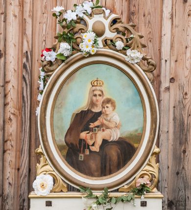 Zdjęcie nr 2: Feretron dwustronny z przedstawieniami Matki Boskiej Niepokalanie Poczętej oraz Matki Boskiej Szkaplerznej ustawiony na prostopadłościennym, profilowanym, zaopatrzonym w otwory na drążki do noszenia cokole. Obrazy w kształcie owalu, umieszczone w profilowanych ramach. Od dołu i góry zdobione liśćmi rzeźbionego akantu. Na obrazie Matki Boskiej Niepokalanie Poczętej Maria ukazana w centrum kompozycji na tle jasnoszarych chmur w postawie stojącej na półksiężycu, delikatnie zwrócona trzy czwarte w swoją prawą stronę. Ma dłonie złożone na piersi, głowę skierowaną w lewo. Ubrana jest w białą suknię z długimi rękawami, zasłaniającą stopy oraz niebieski płaszcz zarzucony jedną połą na lewe przedramię, drugi koniec płaszcza widoczny jest na dole, po prawej stronie Marii. Pod płaszczem chowa się małe putto. Twarz Marii o delikatnych rysach. Włosy bujne, jasne, długie, opadające na plecy i wzdłuż ramion. Kolorystyka jasna. Po drugiej stronie feretronu obraz Matki Boskiej Szkaplerznej. W centrum kompozycji ukazana Maria w pozycji siedzącej, frontalnie, trzymająca na lewym kolanie Dzieciątko Jezus. Maria i Dzieciątko ze szkaplerzami w dłoniach. Matka Boska ubrana jest w habit i płaszcz karmelitański oraz biały welon na głowie. Dzieciątko zwrócone w trzech czwartych w prawo, ze wzrokiem skierowanym na widza. Ubrane w białą, krótka sukienkę. Twarze o delikatnych rysach, idealizowane. Na głowie Marii korona w typie zamkniętym, zwieńczona krzyżykiem. W oddali widoczny pejzaż górski, powyżej błękitne niebo. Kolorystyka ciepła, stonowana. Struktura feretronu polichromowana na biało, detale, ornamentyka i profilowania złocone.
