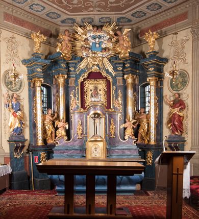 Zdjęcie nr 24: Nastawa ołtarzowa architektoniczna, trójosiowa, jednokondygnacyjna, ustawiona na wysokim cokole. Ołtarz wolnostojący, odsunięty od retabulum, drewniany, sarkofagowy z drewnianą mensą, na nim ustawione tabernakulum. Nastawa opasująca trójbocznie zamknięte prezbiterium, osie wydzielone czterema parami pilastrów o trzonach dekorowanych rocaille'em, przed którymi stoją cztery kolumny o kapitelach kompozytowych i trzonach z dekoracją geometryczną, dźwigających imposty. W polu środkowym nisza w formie stojącego prostokąta zamkniętego łukiem segmentowym nadwieszonym z uskokiem, ujęta profilowaną ramą dekorowaną rocaille'em z dużo mniejszym obrazem Matki Boskiej Nieustającej Pomocy w osobnej ramie na czerwonym tle. Osie boczne przeprute oknami w formie stojącego prostokąta zamkniętego łukiem segmentowym, na ich tle stoją figury aniołów. Nad polem głównym medalion z obłoków i uskrzydlonych główek anielskich z gołębicą Ducha Świętego w glorii promienistej. Na belkowaniu, na osi wewnętrznych impostów dwa siedzące na obłokach putta, na osi zewnętrznych rokokowe wazony. Nastawa flankowana dwoma przyściennymi cokołami, na których stoją figury św. Piotra i św. Pawła. Struktura architektoniczna polichromowana na sposób imitujący niebiesko-białą marmoryzację, trzony pilastrów fioletową. Ornamentyka, profile, kolumny i gloria pozłacane i posrebrzane. 
 





