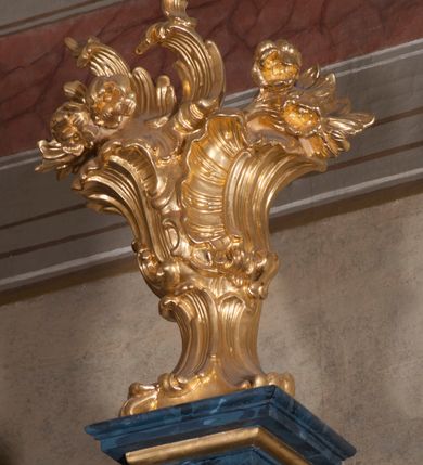 Zdjęcie nr 25: Nastawa ołtarzowa architektoniczna, trójosiowa, jednokondygnacyjna, ustawiona na wysokim cokole. Ołtarz wolnostojący, odsunięty od retabulum, drewniany, sarkofagowy z drewnianą mensą, na nim ustawione tabernakulum. Nastawa opasująca trójbocznie zamknięte prezbiterium, osie wydzielone czterema parami pilastrów o trzonach dekorowanych rocaille&#039;em, przed którymi stoją cztery kolumny o kapitelach kompozytowych i trzonach z dekoracją geometryczną, dźwigających imposty. W polu środkowym nisza w formie stojącego prostokąta zamkniętego łukiem segmentowym nadwieszonym z uskokiem, ujęta profilowaną ramą dekorowaną rocaille&#039;em z dużo mniejszym obrazem Matki Boskiej Nieustającej Pomocy w osobnej ramie na czerwonym tle. Osie boczne przeprute oknami w formie stojącego prostokąta zamkniętego łukiem segmentowym, na ich tle stoją figury aniołów. Nad polem głównym medalion z obłoków i uskrzydlonych główek anielskich z gołębicą Ducha Świętego w glorii promienistej. Na belkowaniu, na osi wewnętrznych impostów dwa siedzące na obłokach putta, na osi zewnętrznych rokokowe wazony. Nastawa flankowana dwoma przyściennymi cokołami, na których stoją figury św. Piotra i św. Pawła. Struktura architektoniczna polichromowana na sposób imitujący niebiesko-białą marmoryzację, trzony pilastrów fioletową. Ornamentyka, profile, kolumny i gloria pozłacane i posrebrzane. 
 





