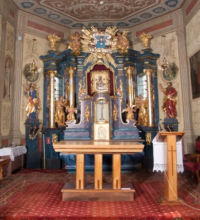 Zdjęcie nr 13: Nastawa ołtarzowa architektoniczna, trójosiowa, jednokondygnacyjna, ustawiona na wysokim cokole. Ołtarz wolnostojący, odsunięty od retabulum, drewniany, sarkofagowy z drewnianą mensą, na nim ustawione tabernakulum. Nastawa opasująca trójbocznie zamknięte prezbiterium, osie wydzielone czterema parami pilastrów o trzonach dekorowanych rocaille'em, przed którymi stoją cztery kolumny o kapitelach kompozytowych i trzonach z dekoracją geometryczną, dźwigających imposty. W polu środkowym nisza w formie stojącego prostokąta zamkniętego łukiem segmentowym nadwieszonym z uskokiem, ujęta profilowaną ramą dekorowaną rocaille'em z dużo mniejszym obrazem Matki Boskiej Nieustającej Pomocy w osobnej ramie na czerwonym tle. Osie boczne przeprute oknami w formie stojącego prostokąta zamkniętego łukiem segmentowym, na ich tle stoją figury aniołów. Nad polem głównym medalion z obłoków i uskrzydlonych główek anielskich z gołębicą Ducha Świętego w glorii promienistej. Na belkowaniu, na osi wewnętrznych impostów dwa siedzące na obłokach putta, na osi zewnętrznych rokokowe wazony. Nastawa flankowana dwoma przyściennymi cokołami, na których stoją figury św. Piotra i św. Pawła. Struktura architektoniczna polichromowana na sposób imitujący niebiesko-białą marmoryzację, trzony pilastrów fioletową. Ornamentyka, profile, kolumny i gloria pozłacane i posrebrzane. 
 





