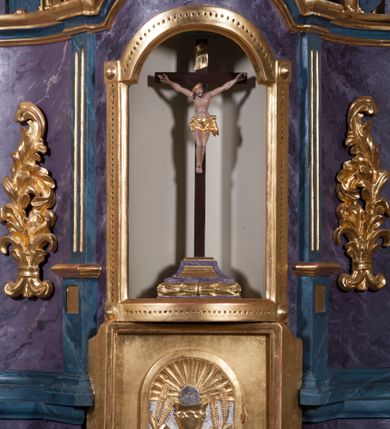 Zdjęcie nr 1: Tabernakulum w formie pozłoconej, prostopadłościennej skrzynki z metalowymi, pozłoconymi i posrebrzonymi drzwiczkami w kształcie stojącego prostokąta zamkniętego łukiem półkolistym z symbolami eucharystycznymi – kielichem z hostią, kłosami zbóż i winnym gronem. Nad tabernakulum tron wystawienia w formie obrotowej niszy o wypukłym licu w architektonicznej obudowie, ujęty spływami wolutowymi, na których znajdują się klęczące figury aniołów, opadającymi na profilowany cokół flankujący tabernakulum. Obrotowa nisza ujęta ramą, flankowana parą pilastrów dźwigających gzyms w formie łuku półkolistego, ujęta ornamentem roślinnym. Pośrodku na gzymsie baranek apokaliptyczny leżący na księdze, na krawędziach gzymsu na cokolikach dwie niewielkie figurki adorujących aniołków. Struktura polichromowana w sposób naśladujący marmoryzację w kolorze fioletowym, z niebieskimi detalami, tabernakulum pozłocone i posrebrzone, ornamentyka i profile pozłocone, figury o naturalnej karnacji i pozłoconych szatach. 

