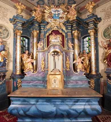 Zdjęcie nr 3: Tabernakulum w formie pozłoconej, prostopadłościennej skrzynki z metalowymi, pozłoconymi i posrebrzonymi drzwiczkami w kształcie stojącego prostokąta zamkniętego łukiem półkolistym z symbolami eucharystycznymi – kielichem z hostią, kłosami zbóż i winnym gronem. Nad tabernakulum tron wystawienia w formie obrotowej niszy o wypukłym licu w architektonicznej obudowie, ujęty spływami wolutowymi, na których znajdują się klęczące figury aniołów, opadającymi na profilowany cokół flankujący tabernakulum. Obrotowa nisza ujęta ramą, flankowana parą pilastrów dźwigających gzyms w formie łuku półkolistego, ujęta ornamentem roślinnym. Pośrodku na gzymsie baranek apokaliptyczny leżący na księdze, na krawędziach gzymsu na cokolikach dwie niewielkie figurki adorujących aniołków. Struktura polichromowana w sposób naśladujący marmoryzację w kolorze fioletowym, z niebieskimi detalami, tabernakulum pozłocone i posrebrzone, ornamentyka i profile pozłocone, figury o naturalnej karnacji i pozłoconych szatach. 

