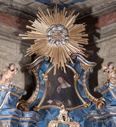 Zdjęcie nr 2: Obraz w formie stojącego prostokąta zamkniętego łukiem segmentowym, o wklęsłych bokach i falistej podstawie. W centrum przedstawiona św. Kinga modląca się przed krucyfiksem. Święta ukazana jest w półpostaci, zwrócona w trzech czwartych w prawo, z głową przechyloną na prawy bark. Jej lewa ręka opuszczona jest w dół, prawa zaś uniesiona. Twarz ma owalną, z wąskimi, migdałowatymi oczami skierowanymi ku górze, dużym nosem i pełnymi ustami. Ubrana jest w czarny habit i czarny płaszcz narzucony na ramiona, na głowie i wokół szyi ma biały kaptur oraz czarny welon z białą podszewką. Przed nią, w lewej części obrazu niewielki stolik, na którym znajduje się krucyfiks, a także korona otwarta oraz księga. Święta ukazana na tle ciemnego, zachmurzonego nieba. 
