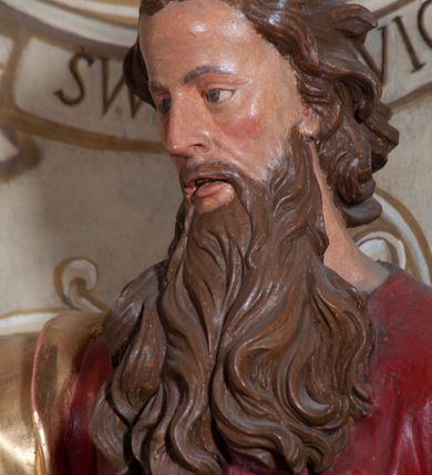 Zdjęcie nr 5: Rzeźba ustawiona na niskiej podstawie przedstawia św. Pawła, który został ukazany frontalnie, w kontrapoście. Lewą ręką podtrzymuje z przodu połę płaszcza i trzyma zamkniętą księgę, prawą unosi do góry z wyciągniętym wskazującym palcem. Twarz pociągła, o blisko siebie usytuowanych oczach, lekko garbatym nosie  i rozchylonych ustach, okolona długą, kręconą brodą opadającą na pierś oraz krótkimi kręconymi włosami. Święty ubrany jest w czerwoną tunikę przylegającą do ciała z szerokimi rękawami, przepasaną w talii o pozłacanym wzorze oraz płaszcz przewiązany diagonalnie przez biodra i zawieszony na prawym przedramieniu. Polichromia ciała naturalistyczna. 


