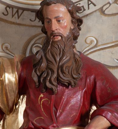 Zdjęcie nr 7: Rzeźba ustawiona na niskiej podstawie przedstawia św. Pawła, który został ukazany frontalnie, w kontrapoście. Lewą ręką podtrzymuje z przodu połę płaszcza i trzyma zamkniętą księgę, prawą unosi do góry z wyciągniętym wskazującym palcem. Twarz pociągła, o blisko siebie usytuowanych oczach, lekko garbatym nosie  i rozchylonych ustach, okolona długą, kręconą brodą opadającą na pierś oraz krótkimi kręconymi włosami. Święty ubrany jest w czerwoną tunikę przylegającą do ciała z szerokimi rękawami, przepasaną w talii o pozłacanym wzorze oraz płaszcz przewiązany diagonalnie przez biodra i zawieszony na prawym przedramieniu. Polichromia ciała naturalistyczna. 


