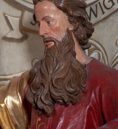 Zdjęcie nr 9: Rzeźba ustawiona na niskiej podstawie przedstawia św. Pawła, który został ukazany frontalnie, w kontrapoście. Lewą ręką podtrzymuje z przodu połę płaszcza i trzyma zamkniętą księgę, prawą unosi do góry z wyciągniętym wskazującym palcem. Twarz pociągła, o blisko siebie usytuowanych oczach, lekko garbatym nosie  i rozchylonych ustach, okolona długą, kręconą brodą opadającą na pierś oraz krótkimi kręconymi włosami. Święty ubrany jest w czerwoną tunikę przylegającą do ciała z szerokimi rękawami, przepasaną w talii o pozłacanym wzorze oraz płaszcz przewiązany diagonalnie przez biodra i zawieszony na prawym przedramieniu. Polichromia ciała naturalistyczna. 


