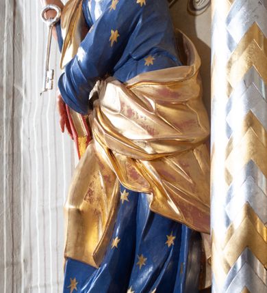 Zdjęcie nr 2: Figura ustawiona na niewielkiej podstawie. Zwrócona jest frontalnie, z uniesioną głową skierowaną w lewo, w kontrapoście, o esowato wygiętej sylwetce. Obie ręce wyciągnięte są w prawo, w lewej święty trzyma czerwoną księgę opartą na prawym udzie, w prawej, uniesionej w górę dłoni trzyma klucze. Twarz ma wychudzoną, o blisko siebie usytuowanych oczach, skierowanych do góry, prostym nosie, otwartych ustach z widocznymi zębami; okoloną krótką, silnie kręconą brodą oraz krótkimi, kręconymi włosami po bokach głowy i nad czołem, na głowie łysina. Święty ubrany jest w przylegającą do ciała niebieską tunikę o szerokich rękawach, z pozłacanymi lamówkami i wzorze z gwiazd. Pozłacany płaszcz jest diagonalnie przewiązany przez korpus, zarzucony na prawe ramię i opadający na prawy bok. Polichromia w partiach ciała naturalistyczna. 
