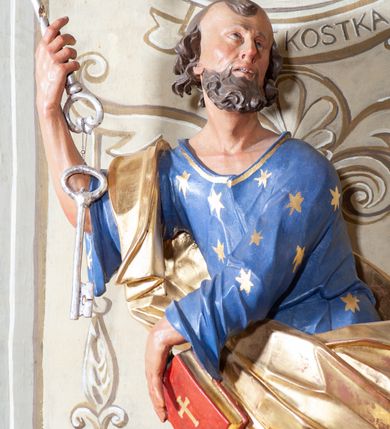 Zdjęcie nr 4: Figura ustawiona na niewielkiej podstawie. Zwrócona jest frontalnie, z uniesioną głową skierowaną w lewo, w kontrapoście, o esowato wygiętej sylwetce. Obie ręce wyciągnięte są w prawo, w lewej święty trzyma czerwoną księgę opartą na prawym udzie, w prawej, uniesionej w górę dłoni trzyma klucze. Twarz ma wychudzoną, o blisko siebie usytuowanych oczach, skierowanych do góry, prostym nosie, otwartych ustach z widocznymi zębami; okoloną krótką, silnie kręconą brodą oraz krótkimi, kręconymi włosami po bokach głowy i nad czołem, na głowie łysina. Święty ubrany jest w przylegającą do ciała niebieską tunikę o szerokich rękawach, z pozłacanymi lamówkami i wzorze z gwiazd. Pozłacany płaszcz jest diagonalnie przewiązany przez korpus, zarzucony na prawe ramię i opadający na prawy bok. Polichromia w partiach ciała naturalistyczna. 
