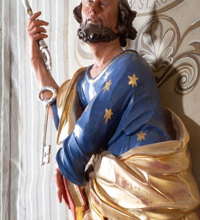Zdjęcie nr 5: Figura ustawiona na niewielkiej podstawie. Zwrócona jest frontalnie, z uniesioną głową skierowaną w lewo, w kontrapoście, o esowato wygiętej sylwetce. Obie ręce wyciągnięte są w prawo, w lewej święty trzyma czerwoną księgę opartą na prawym udzie, w prawej, uniesionej w górę dłoni trzyma klucze. Twarz ma wychudzoną, o blisko siebie usytuowanych oczach, skierowanych do góry, prostym nosie, otwartych ustach z widocznymi zębami; okoloną krótką, silnie kręconą brodą oraz krótkimi, kręconymi włosami po bokach głowy i nad czołem, na głowie łysina. Święty ubrany jest w przylegającą do ciała niebieską tunikę o szerokich rękawach, z pozłacanymi lamówkami i wzorze z gwiazd. Pozłacany płaszcz jest diagonalnie przewiązany przez korpus, zarzucony na prawe ramię i opadający na prawy bok. Polichromia w partiach ciała naturalistyczna. 
