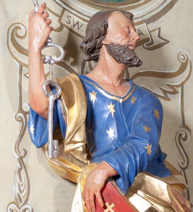 Zdjęcie nr 6: Figura ustawiona na niewielkiej podstawie. Zwrócona jest frontalnie, z uniesioną głową skierowaną w lewo, w kontrapoście, o esowato wygiętej sylwetce. Obie ręce wyciągnięte są w prawo, w lewej święty trzyma czerwoną księgę opartą na prawym udzie, w prawej, uniesionej w górę dłoni trzyma klucze. Twarz ma wychudzoną, o blisko siebie usytuowanych oczach, skierowanych do góry, prostym nosie, otwartych ustach z widocznymi zębami; okoloną krótką, silnie kręconą brodą oraz krótkimi, kręconymi włosami po bokach głowy i nad czołem, na głowie łysina. Święty ubrany jest w przylegającą do ciała niebieską tunikę o szerokich rękawach, z pozłacanymi lamówkami i wzorze z gwiazd. Pozłacany płaszcz jest diagonalnie przewiązany przez korpus, zarzucony na prawe ramię i opadający na prawy bok. Polichromia w partiach ciała naturalistyczna. 
