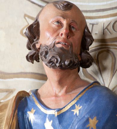 Zdjęcie nr 7: Figura ustawiona na niewielkiej podstawie. Zwrócona jest frontalnie, z uniesioną głową skierowaną w lewo, w kontrapoście, o esowato wygiętej sylwetce. Obie ręce wyciągnięte są w prawo, w lewej święty trzyma czerwoną księgę opartą na prawym udzie, w prawej, uniesionej w górę dłoni trzyma klucze. Twarz ma wychudzoną, o blisko siebie usytuowanych oczach, skierowanych do góry, prostym nosie, otwartych ustach z widocznymi zębami; okoloną krótką, silnie kręconą brodą oraz krótkimi, kręconymi włosami po bokach głowy i nad czołem, na głowie łysina. Święty ubrany jest w przylegającą do ciała niebieską tunikę o szerokich rękawach, z pozłacanymi lamówkami i wzorze z gwiazd. Pozłacany płaszcz jest diagonalnie przewiązany przez korpus, zarzucony na prawe ramię i opadający na prawy bok. Polichromia w partiach ciała naturalistyczna. 
