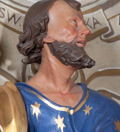 Zdjęcie nr 8: Figura ustawiona na niewielkiej podstawie. Zwrócona jest frontalnie, z uniesioną głową skierowaną w lewo, w kontrapoście, o esowato wygiętej sylwetce. Obie ręce wyciągnięte są w prawo, w lewej święty trzyma czerwoną księgę opartą na prawym udzie, w prawej, uniesionej w górę dłoni trzyma klucze. Twarz ma wychudzoną, o blisko siebie usytuowanych oczach, skierowanych do góry, prostym nosie, otwartych ustach z widocznymi zębami; okoloną krótką, silnie kręconą brodą oraz krótkimi, kręconymi włosami po bokach głowy i nad czołem, na głowie łysina. Święty ubrany jest w przylegającą do ciała niebieską tunikę o szerokich rękawach, z pozłacanymi lamówkami i wzorze z gwiazd. Pozłacany płaszcz jest diagonalnie przewiązany przez korpus, zarzucony na prawe ramię i opadający na prawy bok. Polichromia w partiach ciała naturalistyczna. 
