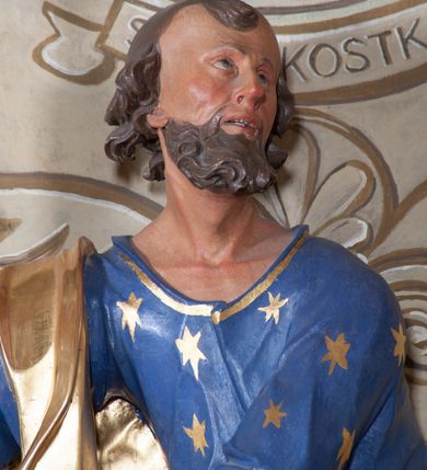 Zdjęcie nr 9: Figura ustawiona na niewielkiej podstawie. Zwrócona jest frontalnie, z uniesioną głową skierowaną w lewo, w kontrapoście, o esowato wygiętej sylwetce. Obie ręce wyciągnięte są w prawo, w lewej święty trzyma czerwoną księgę opartą na prawym udzie, w prawej, uniesionej w górę dłoni trzyma klucze. Twarz ma wychudzoną, o blisko siebie usytuowanych oczach, skierowanych do góry, prostym nosie, otwartych ustach z widocznymi zębami; okoloną krótką, silnie kręconą brodą oraz krótkimi, kręconymi włosami po bokach głowy i nad czołem, na głowie łysina. Święty ubrany jest w przylegającą do ciała niebieską tunikę o szerokich rękawach, z pozłacanymi lamówkami i wzorze z gwiazd. Pozłacany płaszcz jest diagonalnie przewiązany przez korpus, zarzucony na prawe ramię i opadający na prawy bok. Polichromia w partiach ciała naturalistyczna. 
