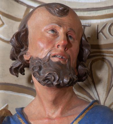 Zdjęcie nr 10: Figura ustawiona na niewielkiej podstawie. Zwrócona jest frontalnie, z uniesioną głową skierowaną w lewo, w kontrapoście, o esowato wygiętej sylwetce. Obie ręce wyciągnięte są w prawo, w lewej święty trzyma czerwoną księgę opartą na prawym udzie, w prawej, uniesionej w górę dłoni trzyma klucze. Twarz ma wychudzoną, o blisko siebie usytuowanych oczach, skierowanych do góry, prostym nosie, otwartych ustach z widocznymi zębami; okoloną krótką, silnie kręconą brodą oraz krótkimi, kręconymi włosami po bokach głowy i nad czołem, na głowie łysina. Święty ubrany jest w przylegającą do ciała niebieską tunikę o szerokich rękawach, z pozłacanymi lamówkami i wzorze z gwiazd. Pozłacany płaszcz jest diagonalnie przewiązany przez korpus, zarzucony na prawe ramię i opadający na prawy bok. Polichromia w partiach ciała naturalistyczna. 
