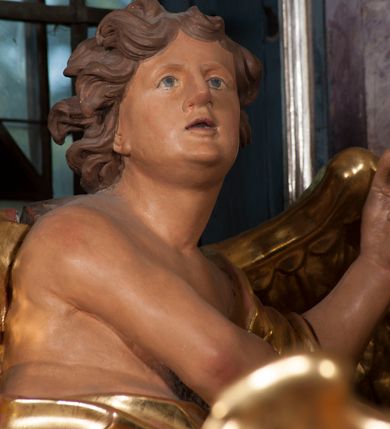 Zdjęcie nr 12: Dwie rzeźby aniołów ustawione są na niewielkich podstawach. Obie figury o układzie ciała w lustrzanym odbiciu względem siebie, zwrócone są frontalnie, w kontrapoście, z głowami skierowanymi w stronę pola głównego ołtarza oraz rękami wyciągniętymi w przeciwną stronę. Twarze mają pociągłe, o młodzieńczych rysach, blisko siebie osadzonych oczach, lekko garbatych nosach, rozchylonych ustach i wystających brodach, okolone ekspresyjnie rozwianymi, krótkimi włosami, rzeźbionymi w pojedyncze pasma. Anioły ubrane są w pozłocone, przylegające do ciała tuniki z jednym rękawem, odsłaniające część torsu i jedną nogę. Obie figury posiadają pozłocone, złożone skrzydła. Polichromia ciała naturalistyczna. 

