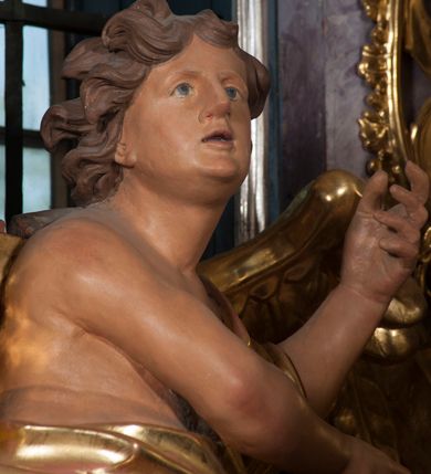 Zdjęcie nr 11: Dwie rzeźby aniołów ustawione są na niewielkich podstawach. Obie figury o układzie ciała w lustrzanym odbiciu względem siebie, zwrócone są frontalnie, w kontrapoście, z głowami skierowanymi w stronę pola głównego ołtarza oraz rękami wyciągniętymi w przeciwną stronę. Twarze mają pociągłe, o młodzieńczych rysach, blisko siebie osadzonych oczach, lekko garbatych nosach, rozchylonych ustach i wystających brodach, okolone ekspresyjnie rozwianymi, krótkimi włosami, rzeźbionymi w pojedyncze pasma. Anioły ubrane są w pozłocone, przylegające do ciała tuniki z jednym rękawem, odsłaniające część torsu i jedną nogę. Obie figury posiadają pozłocone, złożone skrzydła. Polichromia ciała naturalistyczna. 

