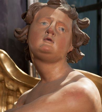 Zdjęcie nr 9: Dwie rzeźby aniołów ustawione są na niewielkich podstawach. Obie figury o układzie ciała w lustrzanym odbiciu względem siebie, zwrócone są frontalnie, w kontrapoście, z głowami skierowanymi w stronę pola głównego ołtarza oraz rękami wyciągniętymi w przeciwną stronę. Twarze mają pociągłe, o młodzieńczych rysach, blisko siebie osadzonych oczach, lekko garbatych nosach, rozchylonych ustach i wystających brodach, okolone ekspresyjnie rozwianymi, krótkimi włosami, rzeźbionymi w pojedyncze pasma. Anioły ubrane są w pozłocone, przylegające do ciała tuniki z jednym rękawem, odsłaniające część torsu i jedną nogę. Obie figury posiadają pozłocone, złożone skrzydła. Polichromia ciała naturalistyczna. 

