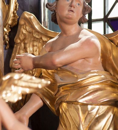 Zdjęcie nr 6: Dwie rzeźby aniołów ustawione są na niewielkich podstawach. Obie figury o układzie ciała w lustrzanym odbiciu względem siebie, zwrócone są frontalnie, w kontrapoście, z głowami skierowanymi w stronę pola głównego ołtarza oraz rękami wyciągniętymi w przeciwną stronę. Twarze mają pociągłe, o młodzieńczych rysach, blisko siebie osadzonych oczach, lekko garbatych nosach, rozchylonych ustach i wystających brodach, okolone ekspresyjnie rozwianymi, krótkimi włosami, rzeźbionymi w pojedyncze pasma. Anioły ubrane są w pozłocone, przylegające do ciała tuniki z jednym rękawem, odsłaniające część torsu i jedną nogę. Obie figury posiadają pozłocone, złożone skrzydła. Polichromia ciała naturalistyczna. 

