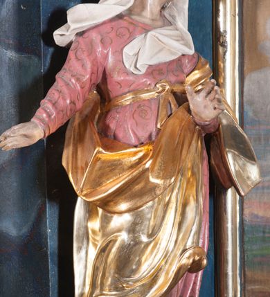 Zdjęcie nr 2: Rzeźba drążona z tyłu, ustawiona na niskiej podstawie z inskrypcją „S(ancta) Anna”. Święta w kontrapoście, zwrócona w trzech czwartych w prawo, z głową skierowaną w lewo. Lewą rękę wyciąga w przód, prawą w bok. Jej twarz ma starcze rysy, lekko garbaty nos, oczy usytuowane blisko siebie oraz pełne, rozchylone usta. Ubrana jest w różową tunikę o pozłoconym wzorze i lamówce, przewiązaną wstęgą w pasie, pozłocony płaszcz zawiązany diagonalnie wokół postaci i zarzucony na lewe ramię, na głowie ma biały welon. Polichromia ciała naturalistyczna. 
