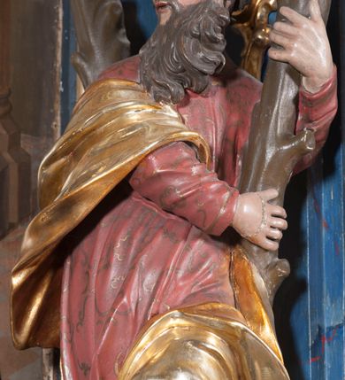 Zdjęcie nr 2: Rzeźba drążona z tyłu, ustawiona na niskiej podstawie. Święty ukazany frontalnie, z lewą nogą opartą na skale, o silnie skręconym w lewo korpusie i głowie skierowanej w przeciwną stronę. Obiema rękami obejmuje ramię stojącego za nim krzyża w kształcie litery „X”. Twarz ma pociągłą, o wysokim czole, lekko garbatym nosie, blisko siebie osadzonych oczach i pełnych, rozchylonych ustach; okolona jest długą, skręconą w pukle brodą, opadającą na pierś oraz krótkimi, kręconymi włosami. Święty ubrany jest w pomarańczowo-różową tunikę o pozłacanym wzorze, przepasaną w talii oraz pozłocony płaszcz przewiązany przez prawe przedramię i opadający na lewą nogę. Polichromia ciała w kolorze naturalistycznym. Na podstawie figury inskrypcja „S(anctus) Andreas”. 
