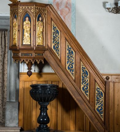 Zdjęcie nr 1: Ambona wisząca, przyścienna, z wejściem od prawej strony w postaci jednobiegowych schodów z balustradą, umieszczona  na północnej ścianie nawy. Kosz na rzucie części ośmioboku, poprzedzony od dołu partią cokołową o mniejszej średnicy, dekorowaną płycinami z ornamentem roślinnym. Dolna krawędź tej podstawy zdobiona maswerkami. Podwieszona do niej szyszka w formie wielokąta o przekroju ośmiobocznym. Ściany kosza dekorowane ostrołukowymi blendami, z płaskorzeźbionymi postaciami czterech ewangelistów; od lewej: Łukasz, Mateusz, Jan i Marek. Postacie są mocno wydłużone, każdy trzyma w rękach księgę, u stóp mają odpowiadające im symbole zwierzęce; każdy jest podpisany. W podłuczach arkadek ornament roślinny, powyżej pas rozetek. W balustradzie schodów romboidalne płyciny wypełnione esowato wygiętą wicią roślinną.
Struktura w kolorze bejcowanego na ciemno drewna; ornamenty, niektóre profile, szaty i atrybuty ewangelistów złocone; nisze malowane na granatowo.  