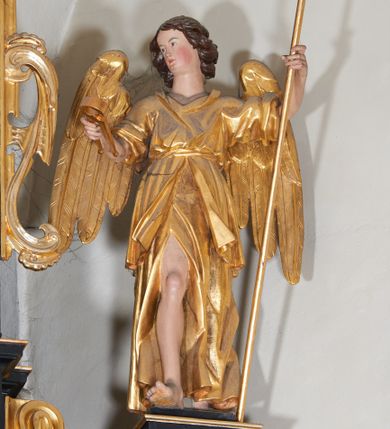 Zdjęcie nr 1: Anioł ukazany w całej postaci, stojący na postumencie po prawej stronie drugiej kondygnacji retabulum. Zwrócony frontalnie, w kontrapoście, z głową skierowaną w stronę ołtarza. Ciężar ciała oparty na lewej nodze, prawa zaś wysunięta w przód. Twarz owalna, osadzona na szerokiej szyi. W twarzy widoczne małe oczy, wąski, delikatnie zadarty nos, niewielkie usta oraz wystająca do przodu, okrągła broda; brązowe, kręcone włosy spływają na kark. W lewej ręce anioł trzyma włócznię z nabitą gąbką, a w prawej obcęgi. Ubrany jest w długą suknię odsłaniającą prawą nogę oraz szatę wierzchnią, przepasaną w talii sznurem; skrzydła złożone. 
Drugi anioł ustawiony jest po drugiej stronie retabulum. Lewą ręką przytrzymuje opartą o ziemię włócznię, a w prawej trzyma młotek. Ubrany jest w długą suknię i tunikę spływające równoległymi fałdami do dołu. Głowa i skrzydła ukształtowane analogicznie jak w przypadku pierwszej postaci. 
W partiach ciała polichromia naturalistyczna, szaty i atrybuty złocone.