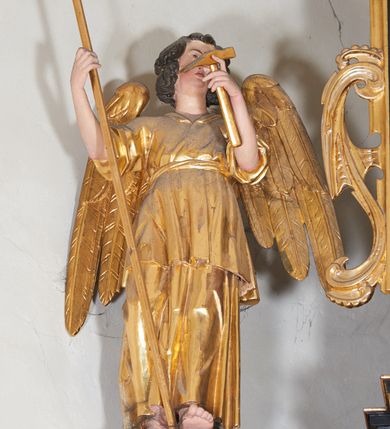 Zdjęcie nr 2: Anioł ukazany w całej postaci, stojący na postumencie po prawej stronie drugiej kondygnacji retabulum. Zwrócony frontalnie, w kontrapoście, z głową skierowaną w stronę ołtarza. Ciężar ciała oparty na lewej nodze, prawa zaś wysunięta w przód. Twarz owalna, osadzona na szerokiej szyi. W twarzy widoczne małe oczy, wąski, delikatnie zadarty nos, niewielkie usta oraz wystająca do przodu, okrągła broda; brązowe, kręcone włosy spływają na kark. W lewej ręce anioł trzyma włócznię z nabitą gąbką, a w prawej obcęgi. Ubrany jest w długą suknię odsłaniającą prawą nogę oraz szatę wierzchnią, przepasaną w talii sznurem; skrzydła złożone. 
Drugi anioł ustawiony jest po drugiej stronie retabulum. Lewą ręką przytrzymuje opartą o ziemię włócznię, a w prawej trzyma młotek. Ubrany jest w długą suknię i tunikę spływające równoległymi fałdami do dołu. Głowa i skrzydła ukształtowane analogicznie jak w przypadku pierwszej postaci. 
W partiach ciała polichromia naturalistyczna, szaty i atrybuty złocone.