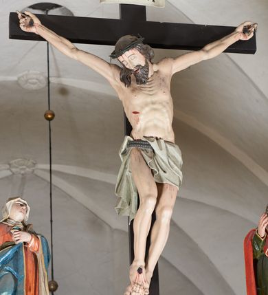 Zdjęcie nr 2: Trójfigurowa grupa Ukrzyżowania, rzeźby pełne. W środku znajduje się krucyfiks z postacią umarłego Chrystusa przybitego trzema gwoździami do krzyża. Chrystus w lekkim zwisie, o szeroko rozłożonych ramionach, bezwładny, głowa opuszczona na prawe ramię. Twarz szczupła, owalna, o prostym nosie, dużych oczach i na wpół otwartych ustach, okolona długim zarostem. Włosy w postaci delikatnych fal opadają na plecy i prawe ramię. Na głowie korona cierniowa. Ciało wychudzone o wyraźnie zarysowanej anatomii. Perizonium w formie tkaniny przewiązanej podwójnym sznurem, odsłaniającej prawe biodro, z dłuższym brzegiem zwisającym po prawej stronie. Draperia gęsto fałdowana, sztywna, o twardych załamaniach materiału. Na szczycie pionowej belki titulus w formie banderoli z napisem „INRI”. Figura Matki Boskiej ustawiona po lewej stronie, zwrócona lekko w stronę krzyża, z głową uniesioną w kierunku Chrystusa. Lewa ręka przyciśnięta do piersi przytrzymuje płaszcz, prawa opuszczona jest wzdłuż ciała. Twarz owalna, niemal bez cech indywidualnych. Ubrana w długą czerwoną suknię, błękitny płaszcz z szarym podbiciem i złoconymi lamówkami, przewiązany diagonalnie przez lewe ramię, opadający na plecy, podtrzymywany na piersi. Na głowie biały welon, opadający na ramiona i plecy. Figura św. Jana Ewangelisty po prawej stronie. Zwrócony delikatnie w stronę krzyża, w kontrapoście, z głową uniesioną w kierunku Chrystusa. Ręce rozpostarte, prawa ugięta i uniesiona w górę, lewa odwiedziona od boku. Twarz młoda, owalna o słabo zindywidualizowanych rysach, włosy w postaci loków sięgają ramion. Święty jest ubrany w ciemnozieloną tunikę przepasaną w talii sznurem oraz czerwony płaszcz ze złoconymi lamówkami, przerzucony diagonalnie przez prawe przedramię i plecy. Matka Boska i św. Jan wykonują podobne gesty; draperie okrywających ich szat są sztywne, ostro załamane. Między figurami Chrystusa i towarzyszącymi mu osobami występuje hierarchia wielkości.