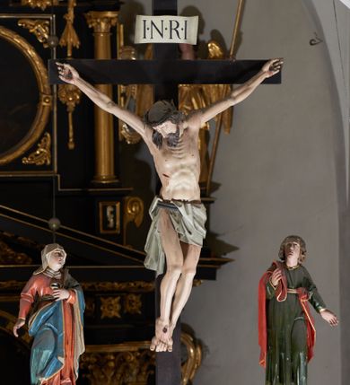 Zdjęcie nr 1: Trójfigurowa grupa Ukrzyżowania, rzeźby pełne. W środku znajduje się krucyfiks z postacią umarłego Chrystusa przybitego trzema gwoździami do krzyża. Chrystus w lekkim zwisie, o szeroko rozłożonych ramionach, bezwładny, głowa opuszczona na prawe ramię. Twarz szczupła, owalna, o prostym nosie, dużych oczach i na wpół otwartych ustach, okolona długim zarostem. Włosy w postaci delikatnych fal opadają na plecy i prawe ramię. Na głowie korona cierniowa. Ciało wychudzone o wyraźnie zarysowanej anatomii. Perizonium w formie tkaniny przewiązanej podwójnym sznurem, odsłaniającej prawe biodro, z dłuższym brzegiem zwisającym po prawej stronie. Draperia gęsto fałdowana, sztywna, o twardych załamaniach materiału. Na szczycie pionowej belki titulus w formie banderoli z napisem „INRI”. Figura Matki Boskiej ustawiona po lewej stronie, zwrócona lekko w stronę krzyża, z głową uniesioną w kierunku Chrystusa. Lewa ręka przyciśnięta do piersi przytrzymuje płaszcz, prawa opuszczona jest wzdłuż ciała. Twarz owalna, niemal bez cech indywidualnych. Ubrana w długą czerwoną suknię, błękitny płaszcz z szarym podbiciem i złoconymi lamówkami, przewiązany diagonalnie przez lewe ramię, opadający na plecy, podtrzymywany na piersi. Na głowie biały welon, opadający na ramiona i plecy. Figura św. Jana Ewangelisty po prawej stronie. Zwrócony delikatnie w stronę krzyża, w kontrapoście, z głową uniesioną w kierunku Chrystusa. Ręce rozpostarte, prawa ugięta i uniesiona w górę, lewa odwiedziona od boku. Twarz młoda, owalna o słabo zindywidualizowanych rysach, włosy w postaci loków sięgają ramion. Święty jest ubrany w ciemnozieloną tunikę przepasaną w talii sznurem oraz czerwony płaszcz ze złoconymi lamówkami, przerzucony diagonalnie przez prawe przedramię i plecy. Matka Boska i św. Jan wykonują podobne gesty; draperie okrywających ich szat są sztywne, ostro załamane. Między figurami Chrystusa i towarzyszącymi mu osobami występuje hierarchia wielkości.