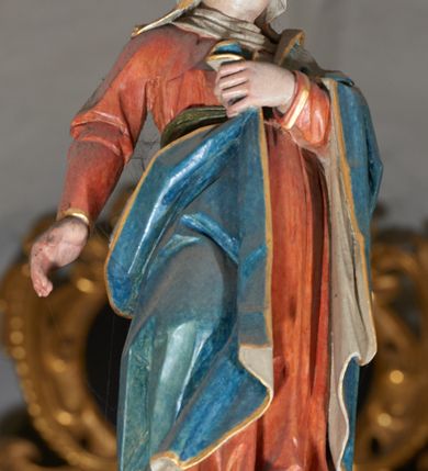 Zdjęcie nr 5: Trójfigurowa grupa Ukrzyżowania, rzeźby pełne. W środku znajduje się krucyfiks z postacią umarłego Chrystusa przybitego trzema gwoździami do krzyża. Chrystus w lekkim zwisie, o szeroko rozłożonych ramionach, bezwładny, głowa opuszczona na prawe ramię. Twarz szczupła, owalna, o prostym nosie, dużych oczach i na wpół otwartych ustach, okolona długim zarostem. Włosy w postaci delikatnych fal opadają na plecy i prawe ramię. Na głowie korona cierniowa. Ciało wychudzone o wyraźnie zarysowanej anatomii. Perizonium w formie tkaniny przewiązanej podwójnym sznurem, odsłaniającej prawe biodro, z dłuższym brzegiem zwisającym po prawej stronie. Draperia gęsto fałdowana, sztywna, o twardych załamaniach materiału. Na szczycie pionowej belki titulus w formie banderoli z napisem „INRI”. Figura Matki Boskiej ustawiona po lewej stronie, zwrócona lekko w stronę krzyża, z głową uniesioną w kierunku Chrystusa. Lewa ręka przyciśnięta do piersi przytrzymuje płaszcz, prawa opuszczona jest wzdłuż ciała. Twarz owalna, niemal bez cech indywidualnych. Ubrana w długą czerwoną suknię, błękitny płaszcz z szarym podbiciem i złoconymi lamówkami, przewiązany diagonalnie przez lewe ramię, opadający na plecy, podtrzymywany na piersi. Na głowie biały welon, opadający na ramiona i plecy. Figura św. Jana Ewangelisty po prawej stronie. Zwrócony delikatnie w stronę krzyża, w kontrapoście, z głową uniesioną w kierunku Chrystusa. Ręce rozpostarte, prawa ugięta i uniesiona w górę, lewa odwiedziona od boku. Twarz młoda, owalna o słabo zindywidualizowanych rysach, włosy w postaci loków sięgają ramion. Święty jest ubrany w ciemnozieloną tunikę przepasaną w talii sznurem oraz czerwony płaszcz ze złoconymi lamówkami, przerzucony diagonalnie przez prawe przedramię i plecy. Matka Boska i św. Jan wykonują podobne gesty; draperie okrywających ich szat są sztywne, ostro załamane. Między figurami Chrystusa i towarzyszącymi mu osobami występuje hierarchia wielkości.